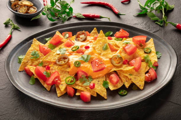 Mexican nachos with a cheese sauce, chili and jalapeno peppers, tomatoes, and cilantro leaves Mexican nachos with a cheese sauce, chili and jalapeno peppers, tomatoes, and cilantro on a black background cheese sauce stock pictures, royalty-free photos & images