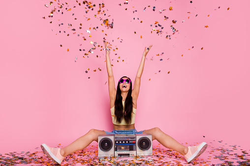 Portrait of her she nice-looking lovely attractive cheerful cheery careless, funky funny girl sitting on floor having fun rising hands up flying decorative elements isolated over pink pastel background