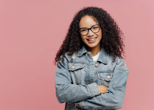 Horizontal shot of mirthful pleasant looking female model wears optical glasses and jean jacket, keeps arms folded over chest, enjoys spare time, nice friendly conversation, poses indoor on pink wall tiro horizontal de alegre agradable aspecto femenino modelo lleva gafas ópticas y chaqueta de jean, mantiene los brazos doblados sobre el pecho, disfruta de tiempo libre, agradable conversación amistosa, poses en el interior en la pared rosa