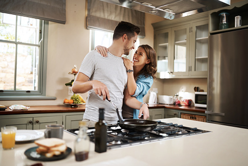 Shot of a young woman embracing her partner while he cooks breakfast