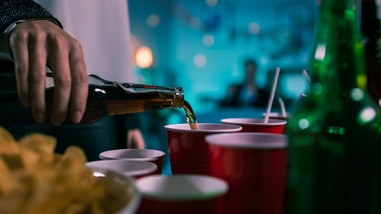 At the Wild Party: Person Pours Drink from a Bottle Into Red Cup. In the Background Blurred Dancing People.