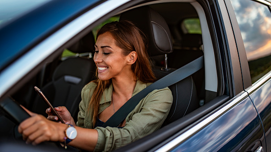 Woman using a navigation app on the smartphone while driving a car. Young woman looking to her smartphone while driving a car. Brown hair happy Woman driving car and texting message on a smartphone during the day.