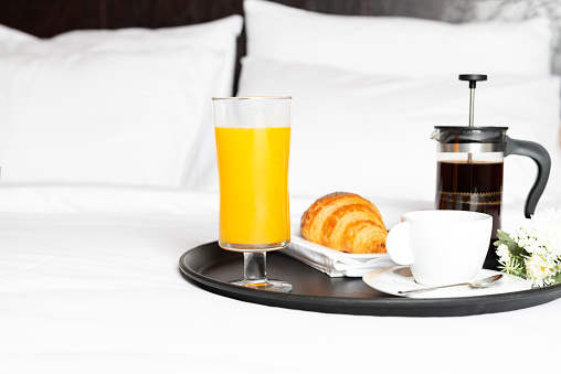 Breakfast with croissant, french press coffee, flowers and newspaper on bed with white sheets.