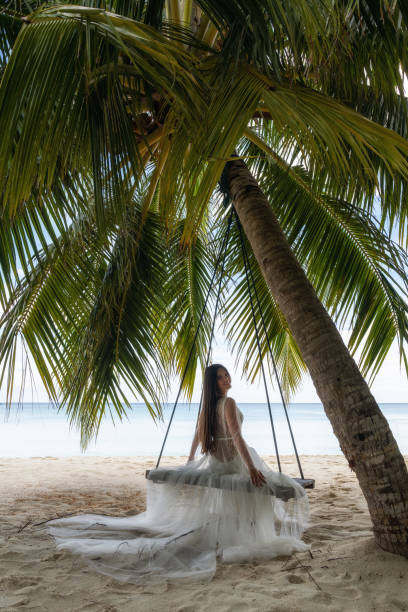 istock A bride in a white dress is riding on a swing under a big palm tree. 1160835224