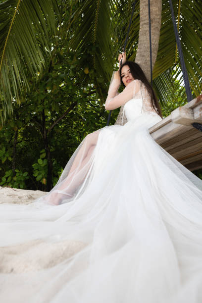 istock A bride in a white dress is riding on a swing under a big palm tree. 1160835225