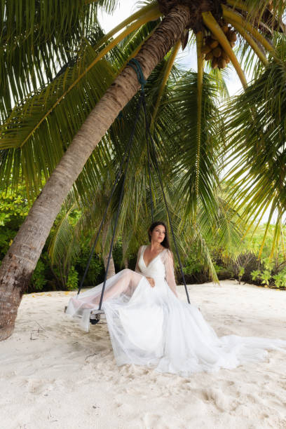 A bride in a white dress is riding on a swing under a big palm tree.
