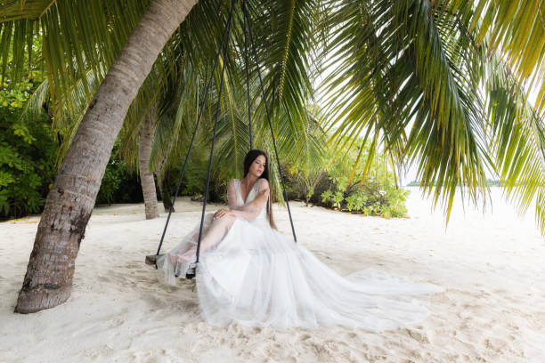 istock A bride in a white dress is riding on a swing under a big palm tree. 1160835255