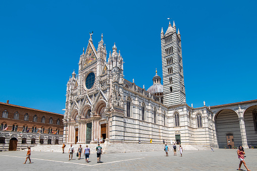 People walking next to the Siena Cathedral at the city of Siena in Tuscany, Italy. It is a medieval church and it was consecrated in the year 1215. It is located in the historic center of Siena and it is an Unesco World Heritage site.