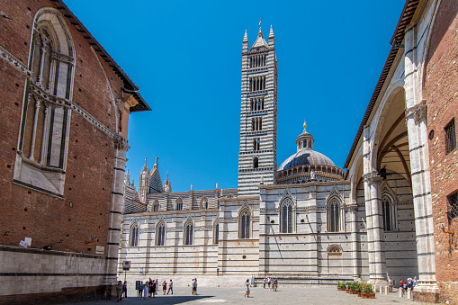 People walking next to the side of Siena Cathedral at the city of Siena in Tuscany, Italy. It is a medieval church and it was consecrated in the year 1215. It is located in the historic center of Siena and it is an Unesco World Heritage site.