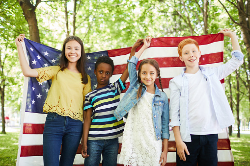 Group of positive multi-ethnic kids in casual outfits standing in forest and holding American flag together while celebrating independence day