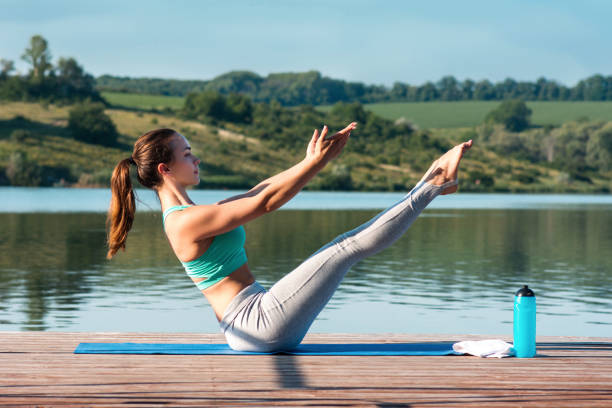 mujer joven deportiva haciendo estiramientos mientras se sienta en el ejercicio de paripurna navasana, equilibrar la postura. hermosa chica atlética practicando yoga en el telón de fondo de la hermosa naturaleza. cálmate, relájate, concepto saludable. - aerobics beautiful bottle body fotografías e imágenes de stock