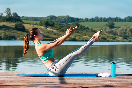 Sporty young woman doing stretching while sitting in Paripurna Navasana exercise, balance posture. Beautiful athletic girl practicing yoga against the backdrop of beautiful nature.Calm down, relax, healthy concept.