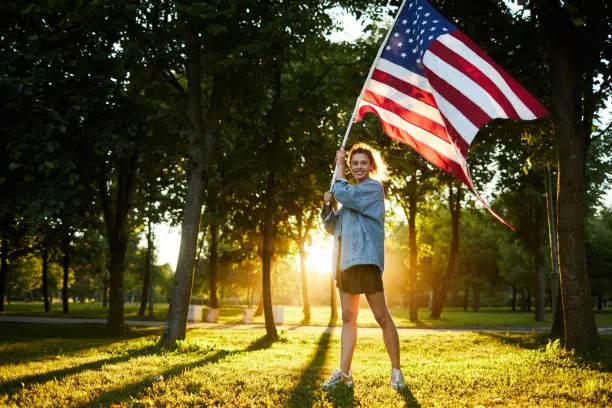 Jolly patriotic young woman in denim jacket standing in forest and holding stars-and-stripes flag