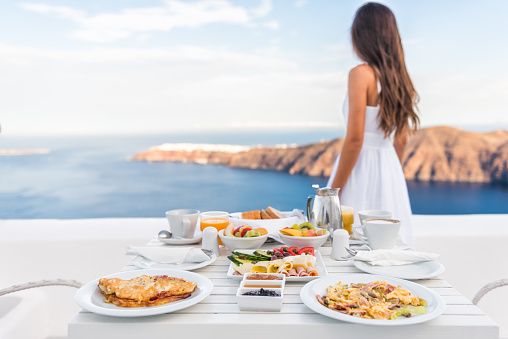 Breakfast table and luxury travel woman on Santorini. Well balanced perfect breakfast table served at resort. Female tourist is looking at beautiful view of sea and caldera enjoying her vacation.