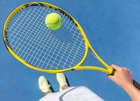Tennis player holding yellow ball on racket grid. Sports female athlete taking a feet selfie showing running shoes on blue hard court. POV closeup of equipment, neon yellow fashion footwear.