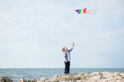 healthy little caucasian boy holding flying colorful kite on autumn sea background