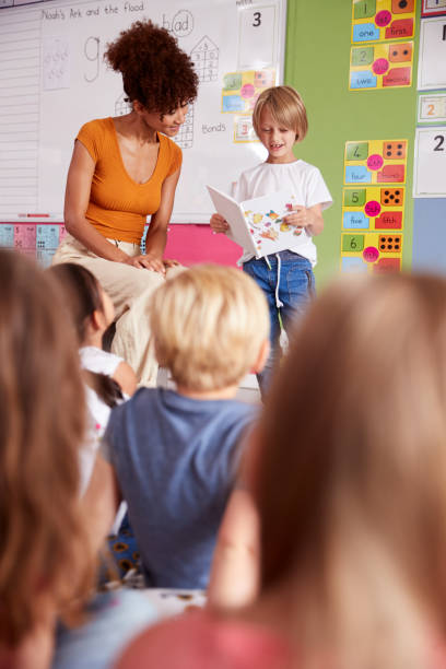 istock Male Pupil In Elementary School Classroom Reading Book To Class With Teacher 1160927466