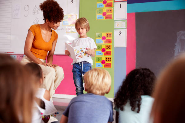 istock Male Pupil In Elementary School Classroom Reading Book To Class With Teacher 1160927620