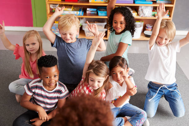 istock Elementary Pupils Raising Hands To Answer Question As Female Teacher Reads Story In Classroom 1160927668