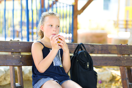 Little girl is eating an apple. Healthy nutrition. Pretty child eating an apple at park , nature outdoors. Teenager pupil enjoying healthy lunch in schoolyard.