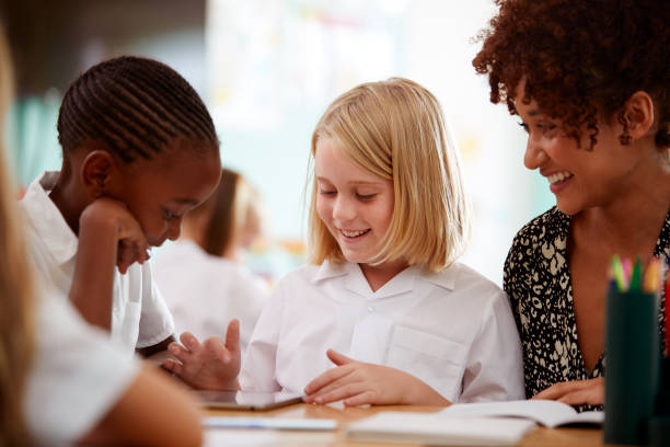 istock Female Teacher With Two Elementary School Pupils Wearing Uniform Using Digital Tablet At Desk 1160931080