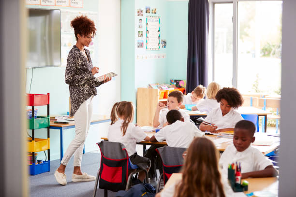 istock Female Teacher With Digital Tablet Teaches Group Of Uniformed Elementary Pupils In School Classroom 1160931174