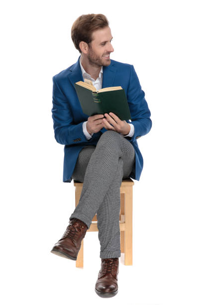 Young man sitting on the couch with legs crossed, wearing glasses and a cardigan, enjoying reading a book in his modern apartment, with a blurred background and a plant in the foreground