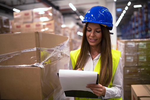 Female factory worker in protective equipment organizing goods arrival. Warehouse inventory manager checking distribution in warehouse storage area. Cardboard boxes in background. Smiling woman at work.