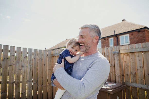 Happy Family Mature father and his baby daughter standing outdoors in the sunshine. The baby girl is embracing her father while looking at the camera. day in the life stock pictures, royalty-free photos & images