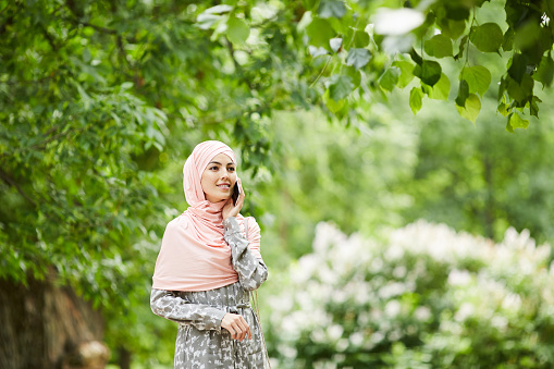 Positive attractive young Muslim woman covering head with hijab standing in summer park and answering call while waiting for friend