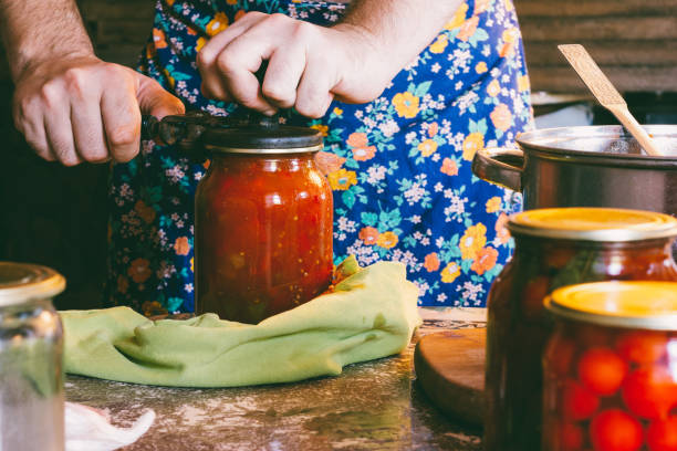 un uomo in grembiule colorato ostrue pomodori e salsa lecho in barattoli di vetro in una fattoria - sugo pronto foto e immagini stock