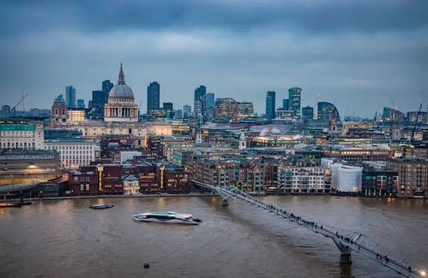 London city skyline with the iconic architecture of Saint Paul's Cathedral and its surroundings, buildings including: Millennium bridge, Thames river, Barbican buildings, illuminated during the blue hour in twilight. Long exposure technique with blurred nautical traffic and tourists/commuters on the foot fridge. Shot with Canon EOS R full frame system and premium lens.