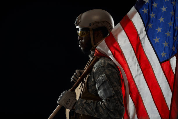 Side view of American ranker with armour confidently standing with weapon and flag on shoulder, looking at camera. African soldier wearing army uniform , preparing for operation.