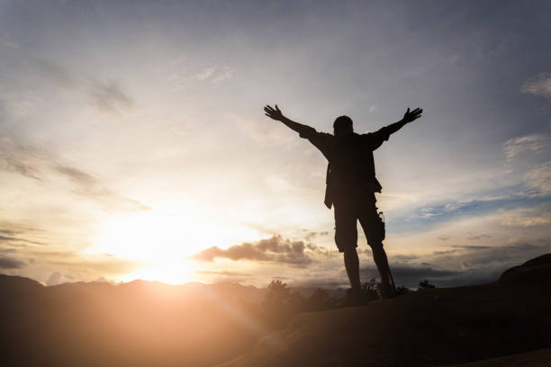 丘の上に立ち、谷の上で日の出を楽しむハイカーのシルエット。 その男は山の上で神に感謝する。 - god freedom arms raised high angle view ストックフォトと画像