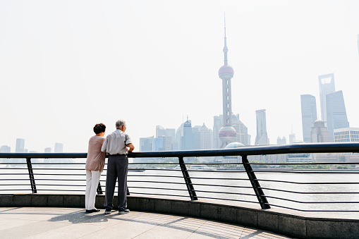 Senior Chinese couple standing at the edge of The Bund and gazing across the Huangpu River at the Pudong skyline.