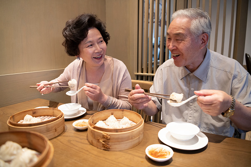 Smiling Chinese seniors talking side by side as delicious dumplings are held between chopsticks and waiting to be consumed.