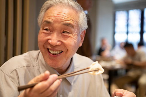 Senior Chinese man holding a dumpling with chopsticks and looking sideways with a mischievous grin before putting it in his mouth.
