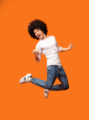 Celebrating success. Happy excited young millennial black with curly hair man jumping on orange background
