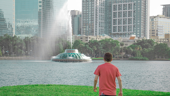 A young man wearing a red t-shirt and water bottle in his back pocket walks out towards a high shooting green fountain in the middle of a beautiful scenic city lake to get a close look.