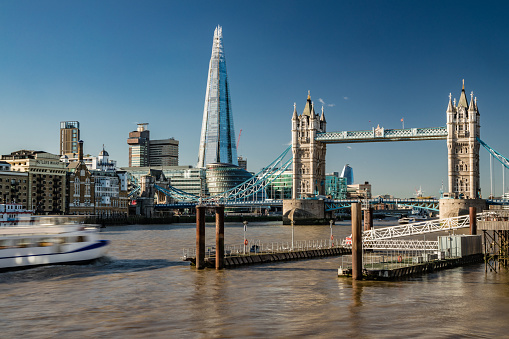 Beautiful colour image of Tower bridge crossing Thames river in London city, including the Shard building and City Hall at the background as photographed from distance on the North bank during the late afternoon hours on a sunny day with clear blue sky. Shot on Canon EOS R full frame system. Long exposure technique to ensure blurred unrecognisable tourist and commuters with blurred traffic including river cruise ferry.