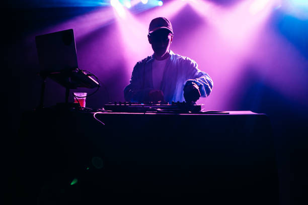 DJ Performing Music Set With Light Display A young African American deejay performs for a crowd at a city night club. Colorful stage lights illuminate the stage behind him. dj photos stock pictures, royalty-free photos & images