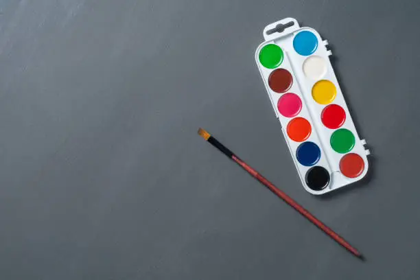 Photo of Box with different colorful watercolor paint near brush with wooden handle on dark blackboard. Copy space for text. Back to school concept