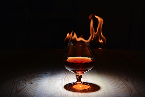 A glass of cognac on a wooden table at dusk on the background of a burning flame.