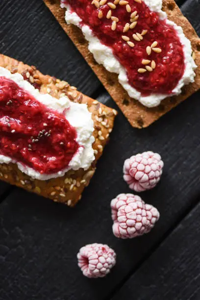 Healthy dessert flat lay. Chia seed jam with creamcheese, flax seeds and raspberries on wholegrain crispbread on black background copy space