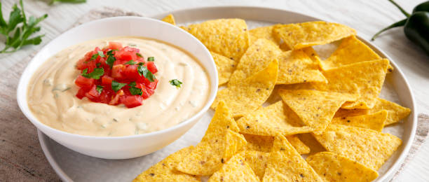 Homemade cheesy dip in a bowl with yellow tortilla chips on a white wooden surface, low angle view. Close-up. Homemade cheesy dip in a bowl with yellow tortilla chips on a white wooden surface, low angle view. Close-up. cheese dip stock pictures, royalty-free photos & images