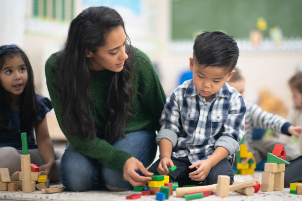 Happy Boy Playing With Blocks A group of preschool kids is indoors in their classroom. They are playing with toy blocks along with their teacher. One boy is very focused on finishing his structure. montessori stock pictures, royalty-free photos & images