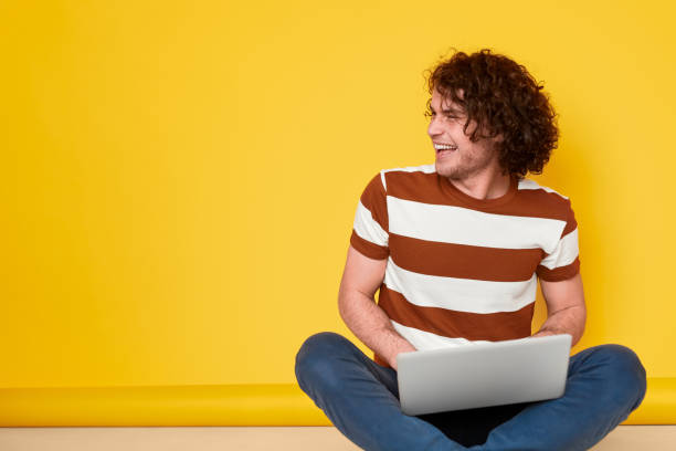 Cheerful male student using laptop Excited young man in casual outfit laughing and looking away while doing homework on laptop against yellow background ecstatic photos stock pictures, royalty-free photos & images