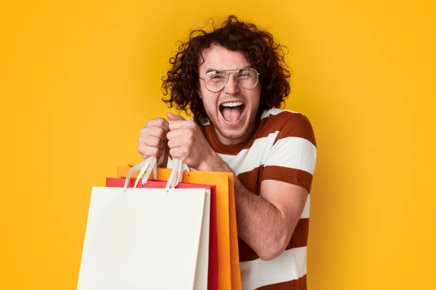 Excited male with shopping bags Funny young male with curly hair and freckles holding bunch of paper bags and screaming in excitement against vivid yellow background shopaholic stock pictures, royalty-free photos & images