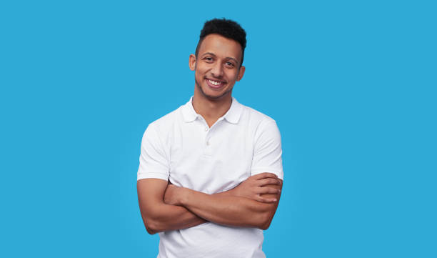 Confident ethnic man smiling for camera Charismatic African American male in white polo shirt keeping arms crossed and smiling while standing against blue background polo shirt stock pictures, royalty-free photos & images