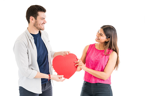 Smiling boyfriend and girlfriend holding red heart shape paper while looking at each other in studio